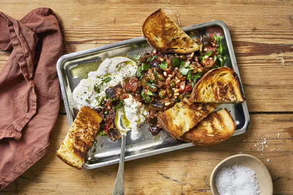 En krämig, vegetarisk midsommarmat med rostade grönsaker, oliver, pinjenötter och vitlöksbröd på ett serveringsfat.