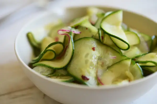 En skål med tunt skivad gurka, perfekt för ett enkelt gurksallad recept. Små röda chiliflakes och lila strimmor av rädisa syns bland gurkskivorna.