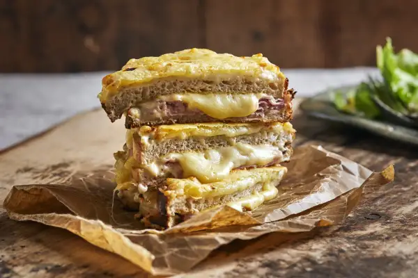En stapel gyllene, smältande grilled cheese-mackor med skinka, serverade på brunt papper.