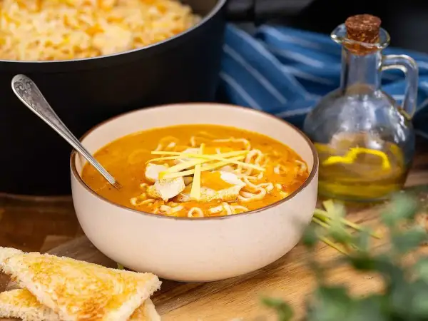 En skål med kycklingsoppa med nudlar, toppad med strimlad kyckling och grönt. Bredvid syns rostat bröd och en flaska olja.