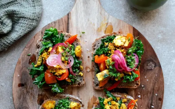 Fyra goda mackor med tofu scramble, grönkål, paprika och rödlök på grovt bröd, serverade på en träskärbräda.