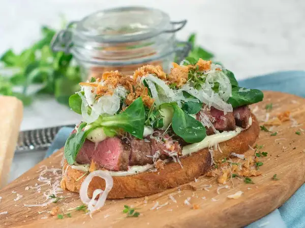 En läcker steak sandwich med saftigt kött, krispig sallad, hyvlad parmesan och friterad lök på rostat bröd.