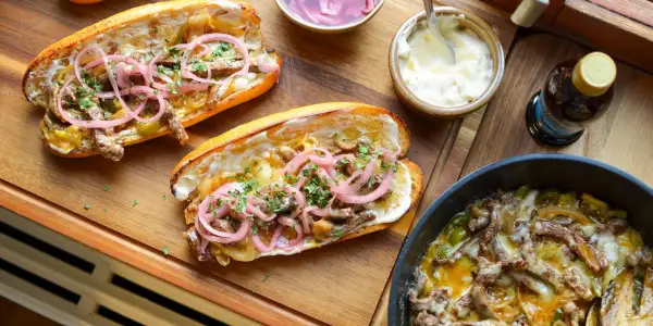 Två saftiga philly cheese steak-mackor med strimlat kött, lök, paprika och toppade med picklad rödlök och persilja. Serveras med majonnäs och en flaska dryck.